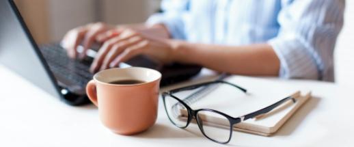 Person working on a laptop with a cup of coffee, notepad, and glasses on the desk